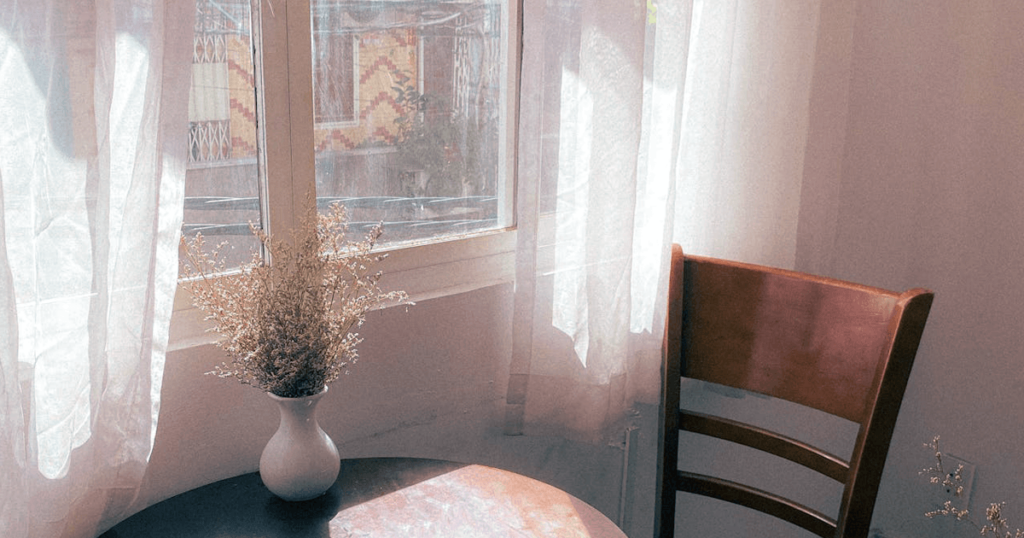 Soft sunlight streams through sheer white curtains, illuminating a cozy corner with a wooden chair and a small round table. A simple white vase holding delicate dried flowers sits on the table, casting subtle shadows. The window offers a glimpse of an exterior wall with a geometric brick pattern, adding warmth and texture to the serene atmosphere.