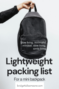 A minimalist black backpack held by a hand against a light background, overlaid with text that reads 'Lightweight packing list for a mini backpack' and phrases promoting slow living and a minimalist mindset from bridgetsbloomzone.com.