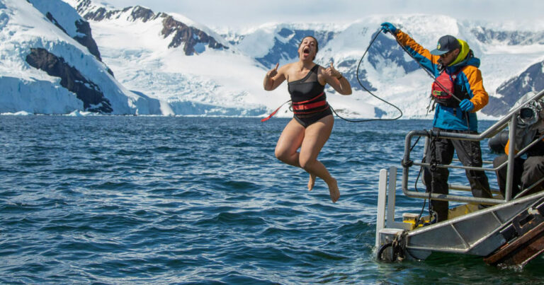 A woman in a black swimsuit takes a daring leap into icy Antarctic waters from a small platform, tethered to a safety line held by an assistant dressed in bright outdoor gear. The scene is set against a breathtaking backdrop of snow-covered mountains and glaciers, with the deep blue ocean reflecting the cold, crisp atmosphere of the polar environment.