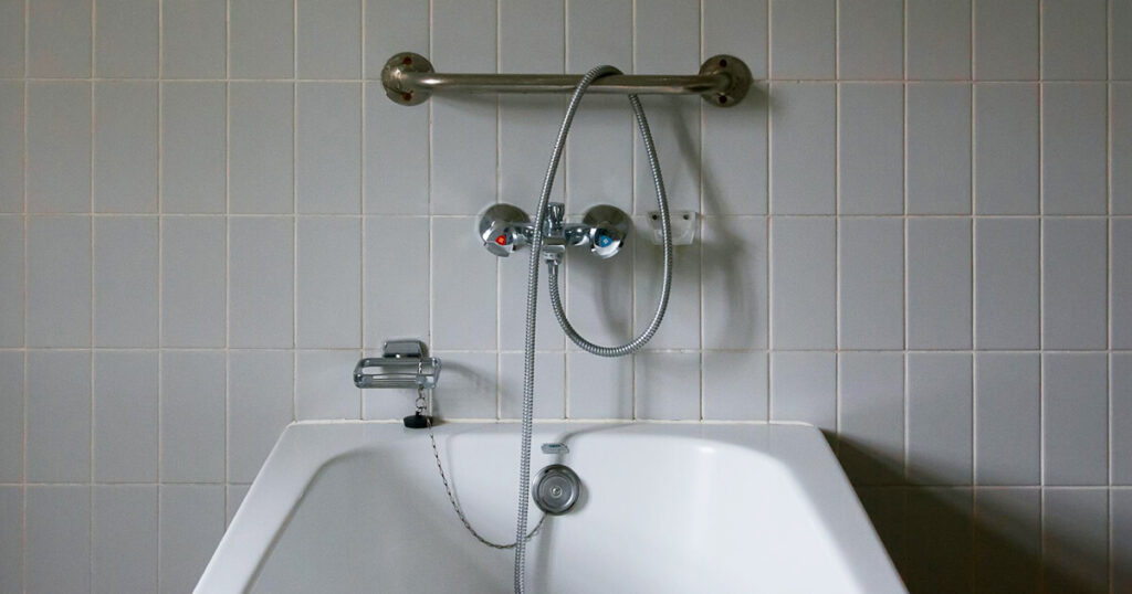 A minimalistic bathroom scene featuring a white tiled wall, a chrome faucet with a handheld showerhead, and a sturdy grab bar mounted above. The clean lines and neutral tones create a functional and understated atmosphere, emphasizing simplicity and the only task you have when taking a shower.