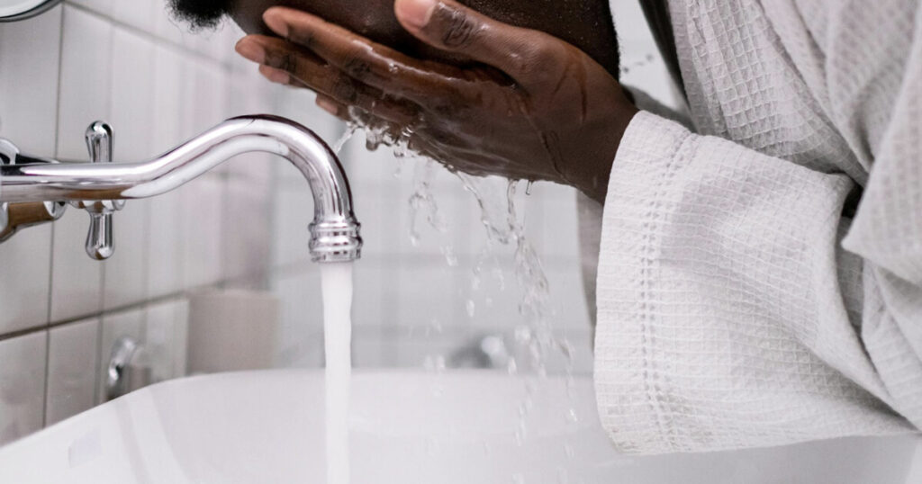 A close-up of a man washing their face with water from a chrome faucet in a bright, tiled bathroom. He is wearing a textured white bathrobe, and water flows gently over their hands and into the sink, creating a refreshing and calming moment. 
