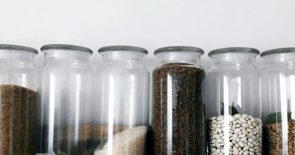 An aesthetically pleasing row of glass jars neatly arranged against a white background. Each jar is filled with pantry essentials such as sugar, grains, coffee grounds, and beans, showcasing an organized and minimalist approach to storage. The clear glass and uniform design create a clean and modern look.