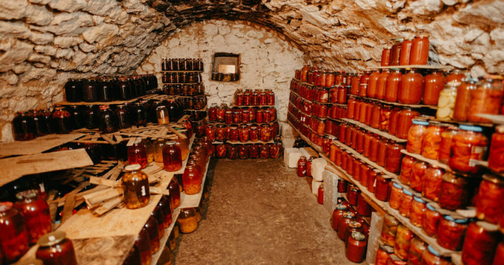 A rustic underground cellar with arched stone walls, filled with neatly arranged shelves of preserved foods in glass jars. The jars contain a variety of vibrant pickled vegetables, jams, and sauces, creating a colorful display against the earthy tones of the stone. The warm lighting enhances the cozy, old-world charm of this traditional storage space.