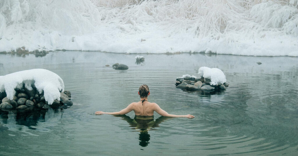 A serene winter scene of a woman immersed in a lake, surrounded by snow-covered rocks and frosted trees. With her back to the camera, she stretches her arms outwards, embracing the tranquil atmosphere. The stillness of the water reflects the peaceful ambiance of the snowy landscape.