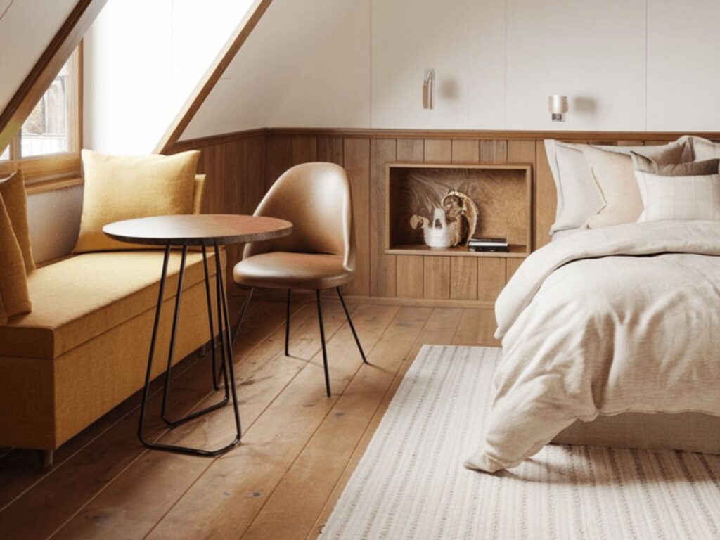 Small attic bedroom with slanted ceiling, built-in bench, round table, warm wood accents, and neutral cozy bedding.