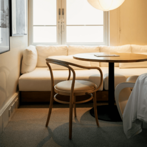 Cozy small dining nook with a round wooden table, a curved wooden chair, and a built-in white sofa by the window, illuminated by soft natural light and a minimalist pendant lamp.
