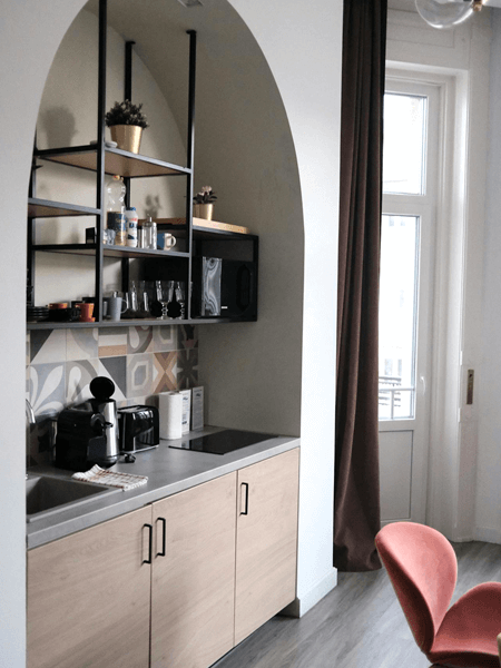 Small kitchenette with light wood cabinets, a gray countertop, open black metal shelving, and patterned backsplash tiles, framed by an arched wall, combining functionality and style in a compact space.