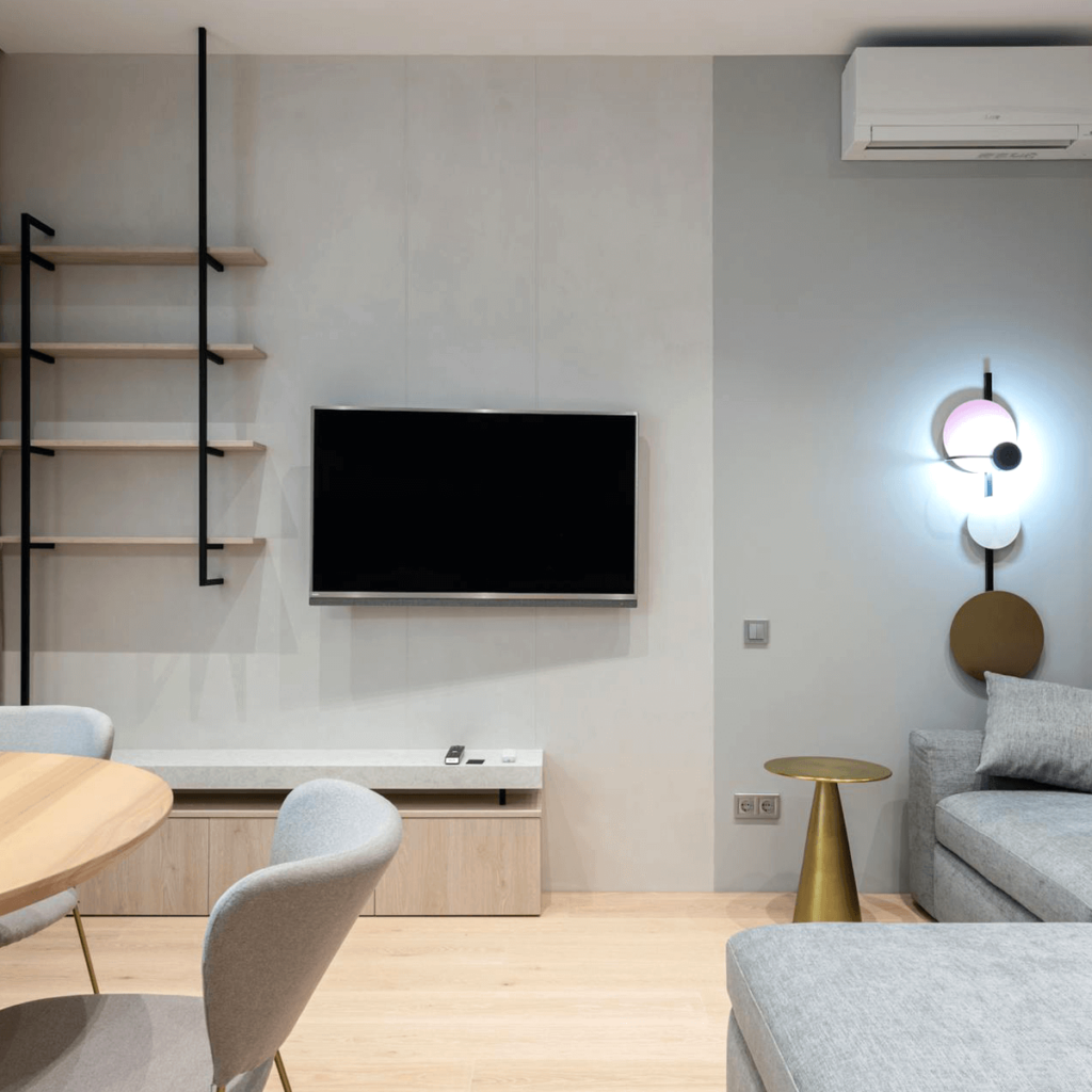 Modern small living room with a wall-mounted TV, light wood storage unit, floating black metal shelves, a gray sofa, a gold side table, and a decorative wall lamp, blending functionality and style in a compact space.