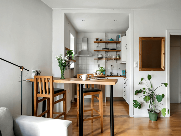 Small open-plan living and kitchen area with a wooden high-top dining table, matching bar stools, white cabinets, open shelving, and decorative touches like a gold-framed mirror and a potted plant, showcasing practical furniture choices for compact spaces.