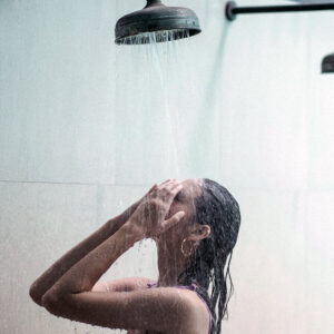 A refreshing shower scene with water cascading from an overhead showerhead onto a person standing beneath it. The minimalist bathroom setting highlights the invigorating power of routines like cold showers to boost energy and focus.