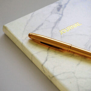 Close-up of a luxurious gold pen resting atop a marble-patterned journal with the word 'JOURNAL' embossed in gold lettering. The elegant design and neutral tones evoke a sense of sophistication, reflection, and the importance of writing as a tool for productivity and achieving your goals.