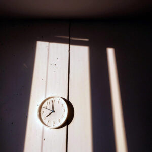 A wall-mounted clock displaying 8:00, illuminated by sunlight streaming through vertical blinds. The interplay of light and shadow creates a serene and reflective atmosphere, symbolizing time management and mindfulness which is key for achieving your goals.