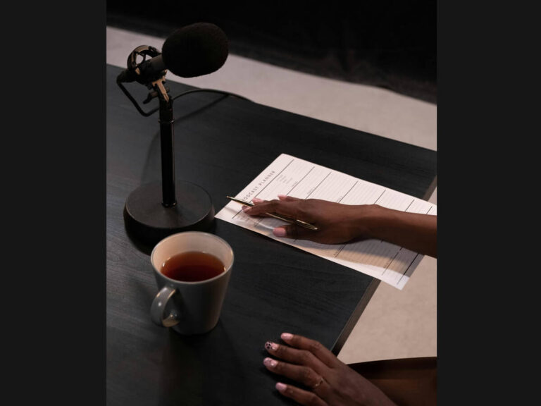 An elegant workspace featuring a black desk with a microphone on a stand, a cup of tea, and a hand holding a pen poised over a structured planning sheet labeled 'Podcast Planning.' The scene conveys focus, organization, and the quiet determination needed for productivity and goal-setting.