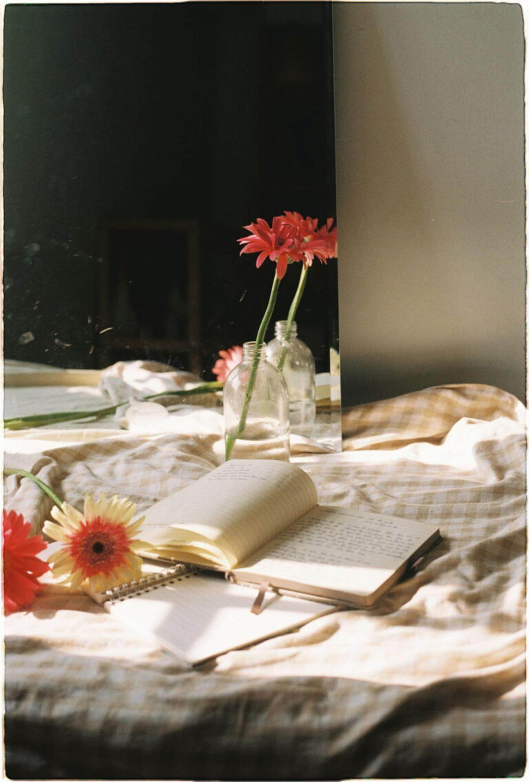 Open journal and notebook with handwritten pages, a pen, and red and yellow gerbera daisies arranged on a soft, sunlit bed with a checkered blanket, next to a glass vase and mirror, evoking a peaceful, minimalist atmosphere that reflects slow living and the calming simplicity of a thoughtfully designed peaceful home.