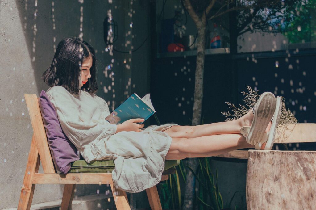 A girl reading outside -girl lounging on a wooden bench outdoors, wearing a light dress and reading a book in dappled sunlight, surrounded by greenery, capturing a tranquil moment of slow, mindful living.