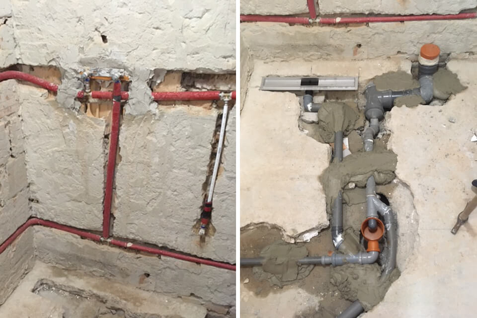 Exposed plumbing installation in a small bathroom under renovation, showing red and silver water pipes embedded in a wall and gray PVC drain pipes set into a concrete floor, illustrating the early stages of bathroom remodeling with visible pipework and unfinished surfaces.