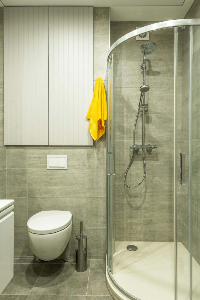 Compact bathroom with gray tiles, a curved glass shower enclosure, wall-mounted toilet, and a yellow towel hanging on the wall.