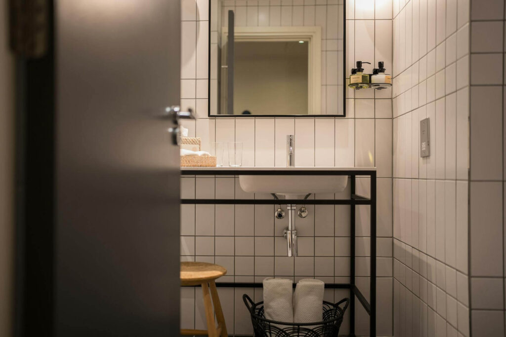 Minimalist small bathroom with white rectangular tiles, a large wall mirror, a modern sink with a black metal frame, and a wire basket holding rolled towels underneath, illustrating how clean lines, open storage, and reflective surfaces can make a compact space feel organized and bright.
