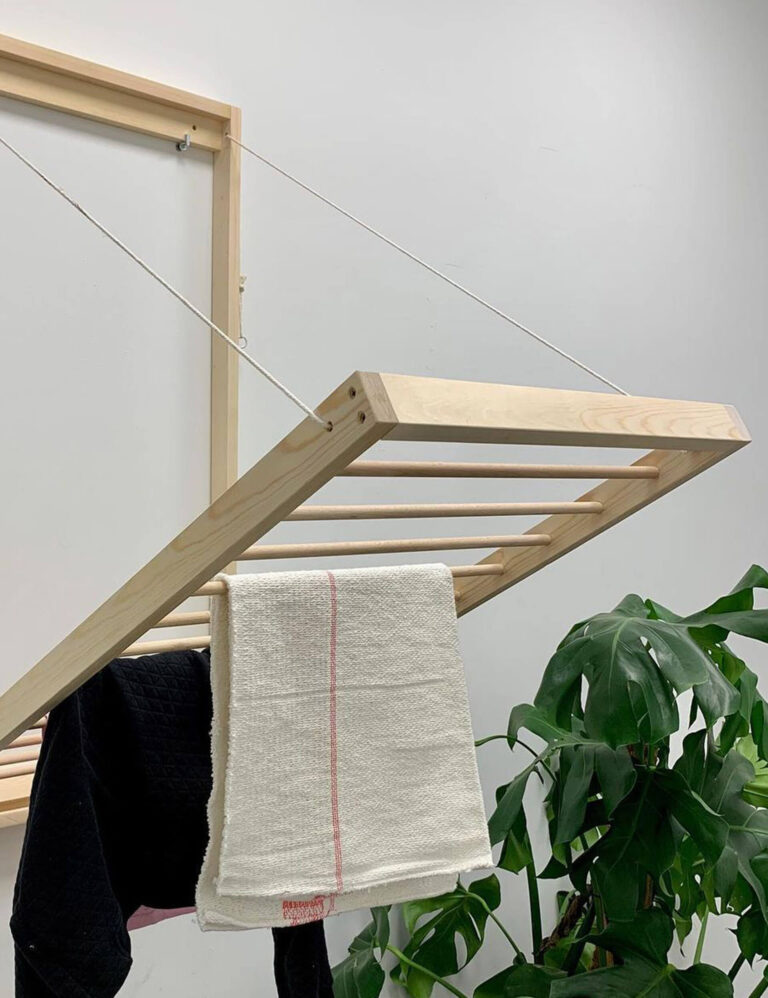 Wall-mounted folding wooden drying rack with towels hanging on it, set against a white wall and a leafy green plant.