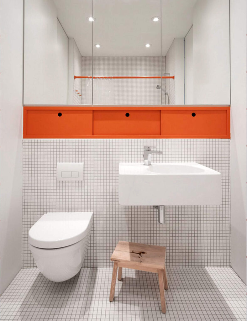 Minimalist bathroom with tiny mosaic tiles, floating toilet and sink, wooden stool, and built-in orange storage cubbies beneath mirrored cabinets.