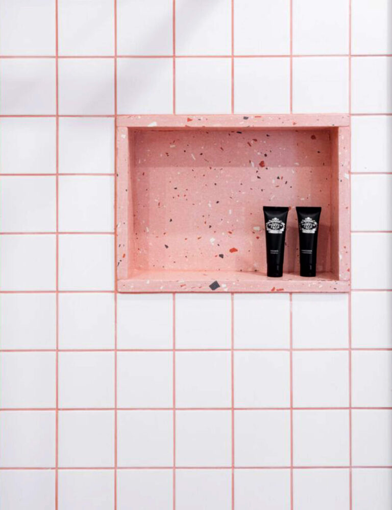 Shower wall with a pink terrazzo built-in niche holding two black toiletry tubes, surrounded by white tiles with pink grout.