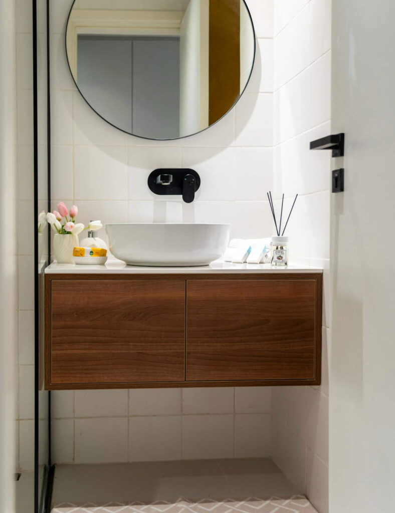 Small bathroom with round mirror and floating wooden vanity under a counter-top sink, accessorized with flowers and scented sticks.