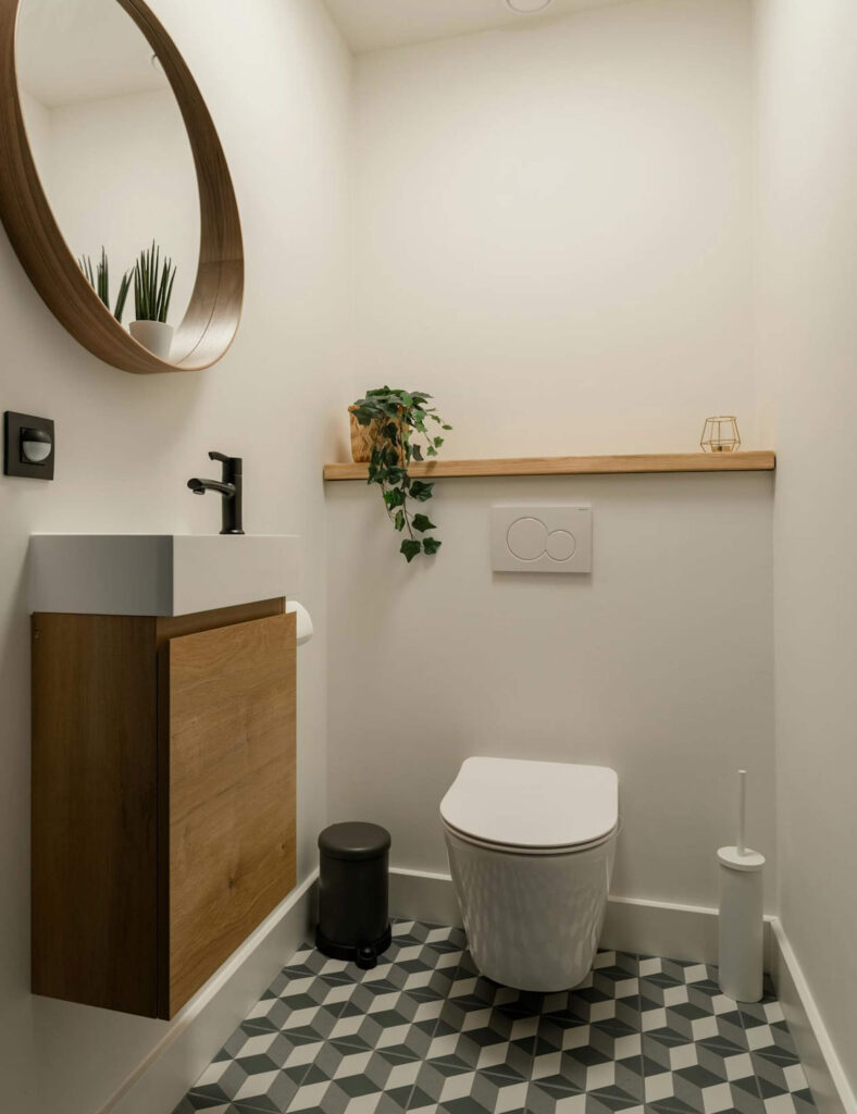 Small bathroom with round mirror and floating wooden vanity under a counter-top sink, accessorized with flowers and scented sticks.