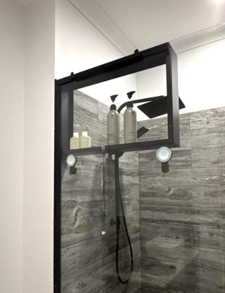 Modern shower with grey wood-look tiles and a black framed glass panel, featuring a shower caddy mounted on the glass holding shampoo and body wash bottles.
