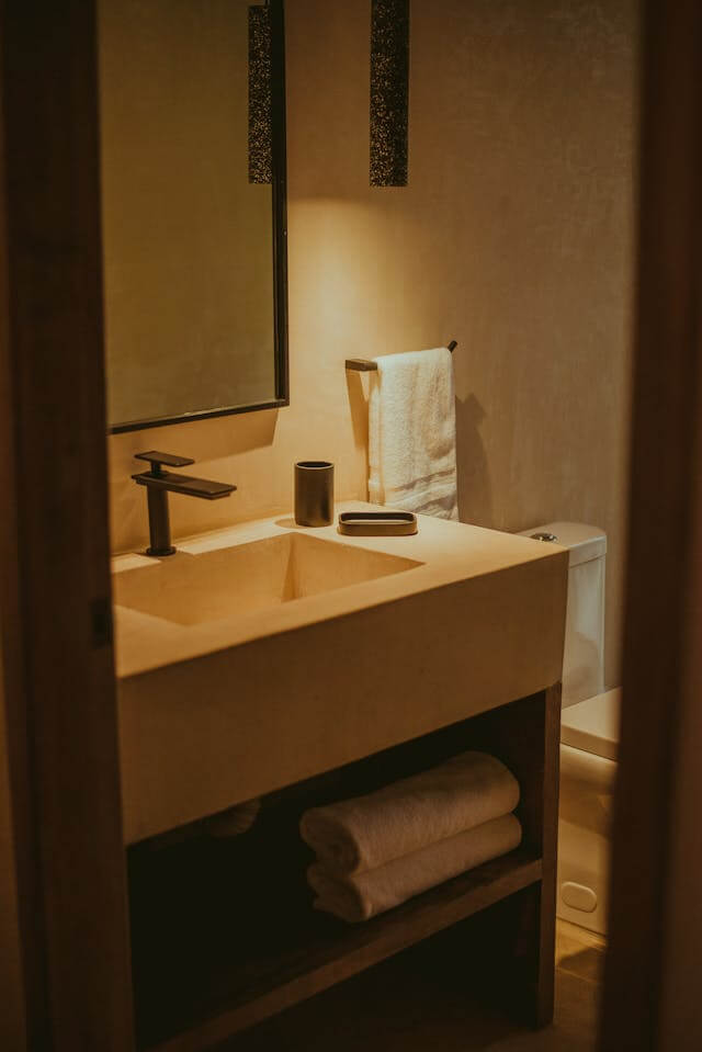 Warmly lit small bathroom featuring a minimalist sink with black fixtures, an open wooden shelf storing rolled towels, and a white towel hanging by the toilet.