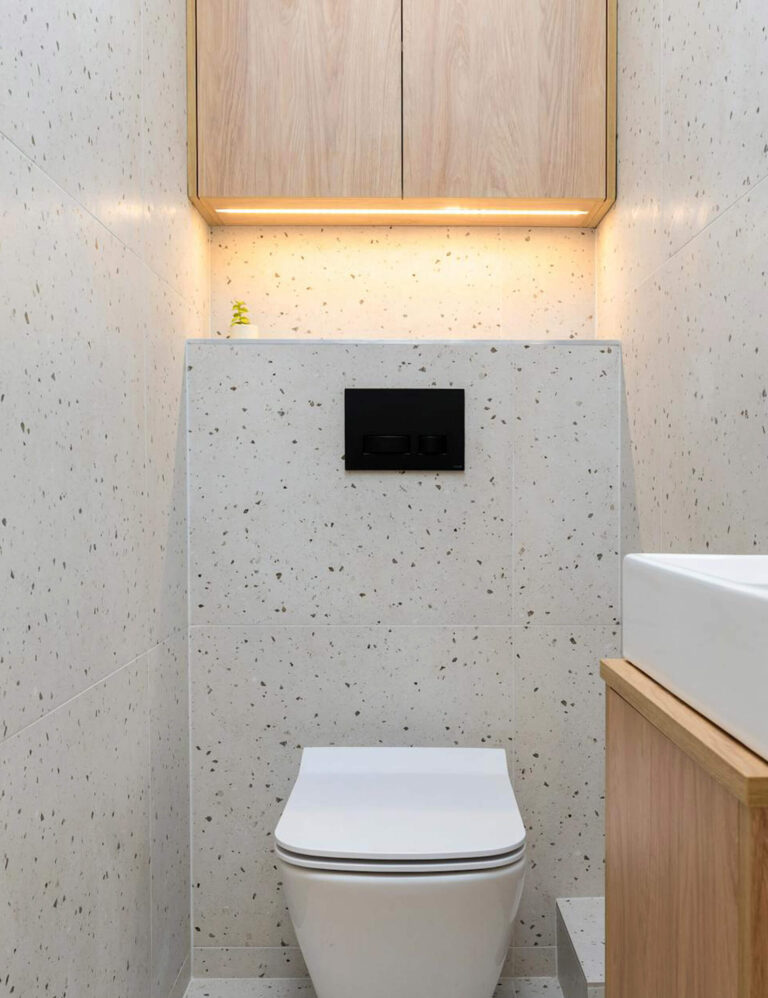 Minimalist bathroom with terrazzo walls and a wall-mounted toilet beneath sleek wooden cabinets with under-cabinet lighting.
