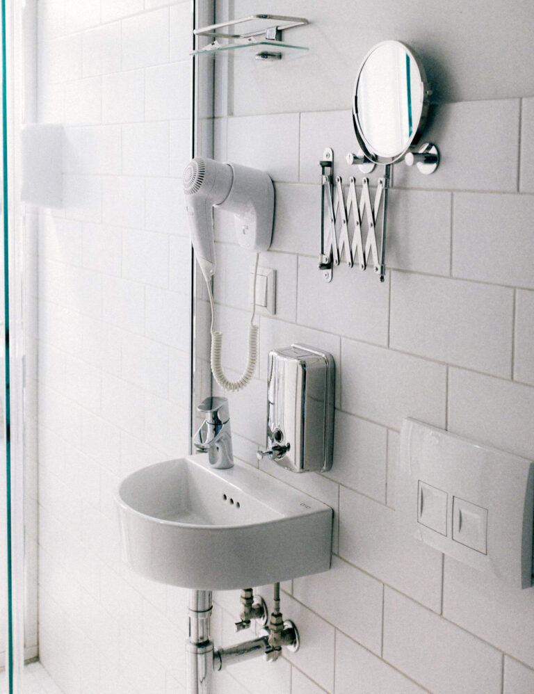 Small bathroom with a wall-mounted sink, hair dryer, soap dispenser, glass shelf, and extendable mirror installed on white tiled walls.