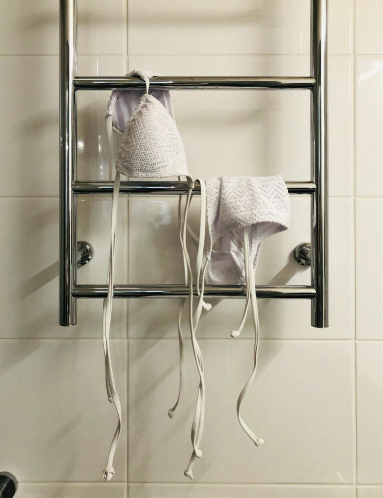 Close-up of a chrome towel rail being used to air dry a white bikini top against tiled bathroom walls.