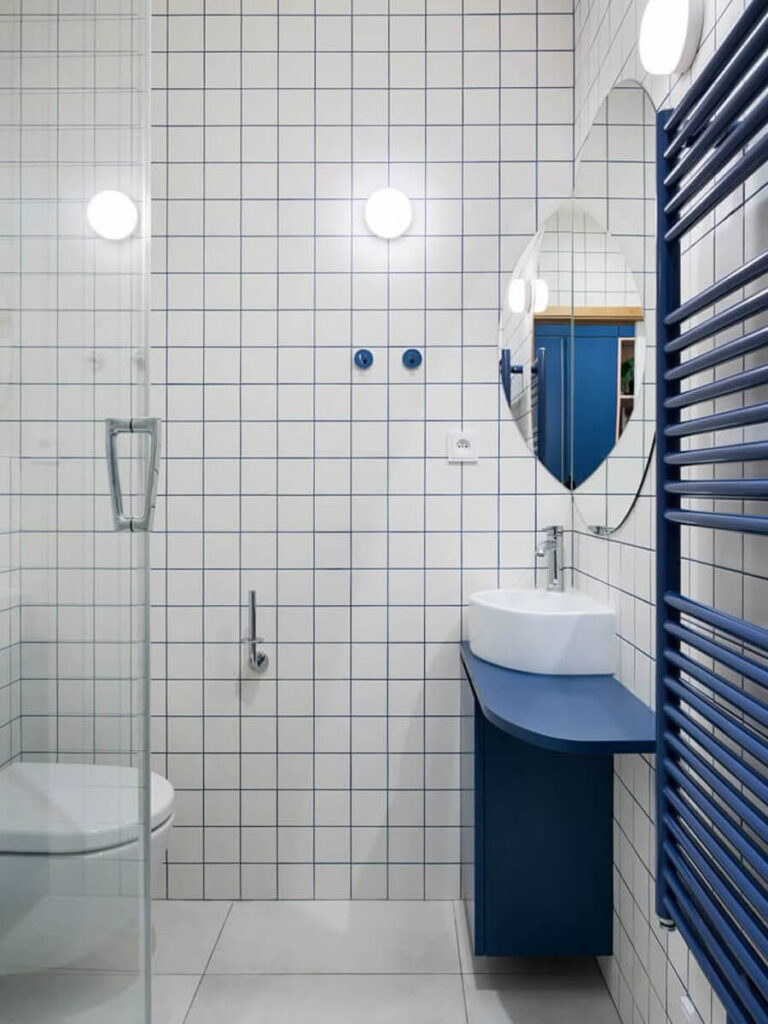 Minimalist bathroom with square white tiles and blue grout, blue towel radiator and vanity, a rounded white sink, oval mirror, soft globe wall lights, and glass shower enclosure for a clean, modern design.