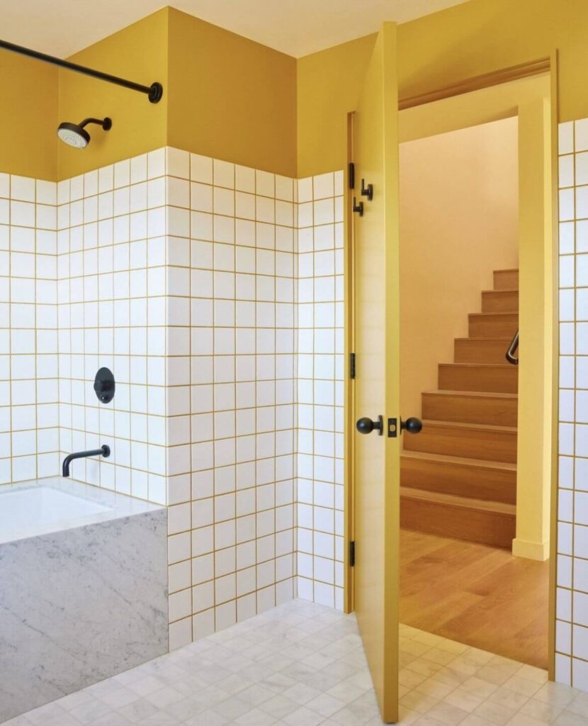 Contemporary bathroom with white subway tiles and yellow grout, a sleek black shower fixture, and marble bathtub surround, adjacent to a mustard yellow wall and door.
