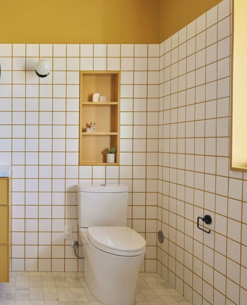 Modern bathroom featuring bold yellow grout between white subway tiles, mustard wall paint and shelving, a white toilet, and marble flooring.