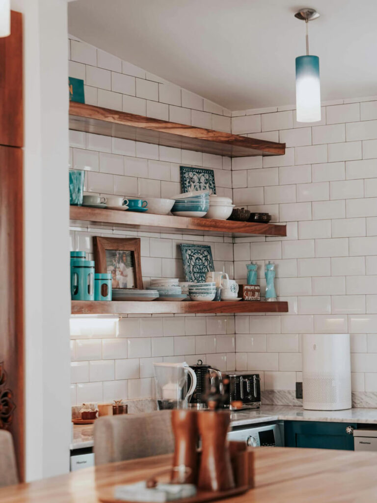 Cozy kitchen with glossy subway backsplash tiles arranged in a running bond pattern, white grout, natural wood open shelving, and turquoise accent dishware for a warm, inviting atmosphere.​​