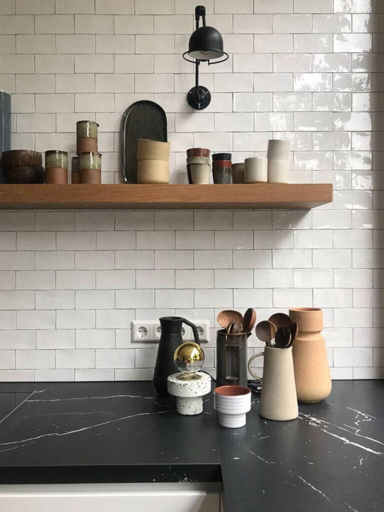 Modern kitchen workspace with glossy white subway tiles laid in running bond, white grout, matte black wall lamp, wood open shelf, and a variety of earth-toned ceramic vessels on a dark countertop.