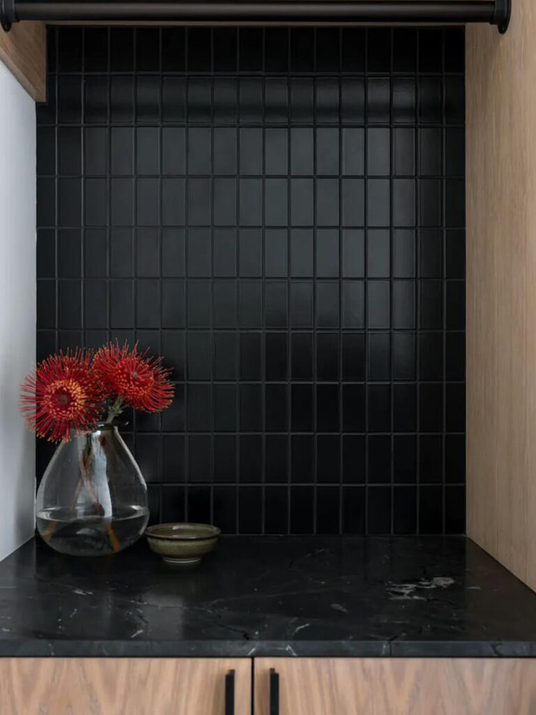 Matte black square tiles set in a vertical grid form a dramatic kitchen backsplash, surrounded by natural wood cabinetry and decorated with a glass vase of red flowers.