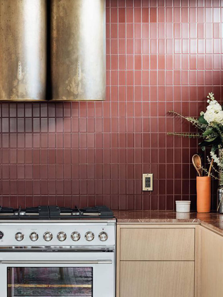 Vertical matte brown tiles form a backsplash in a kitchen, paired with earthy wood cabinetry, brown fixtures, and a pop of vibrant brass vent hood.