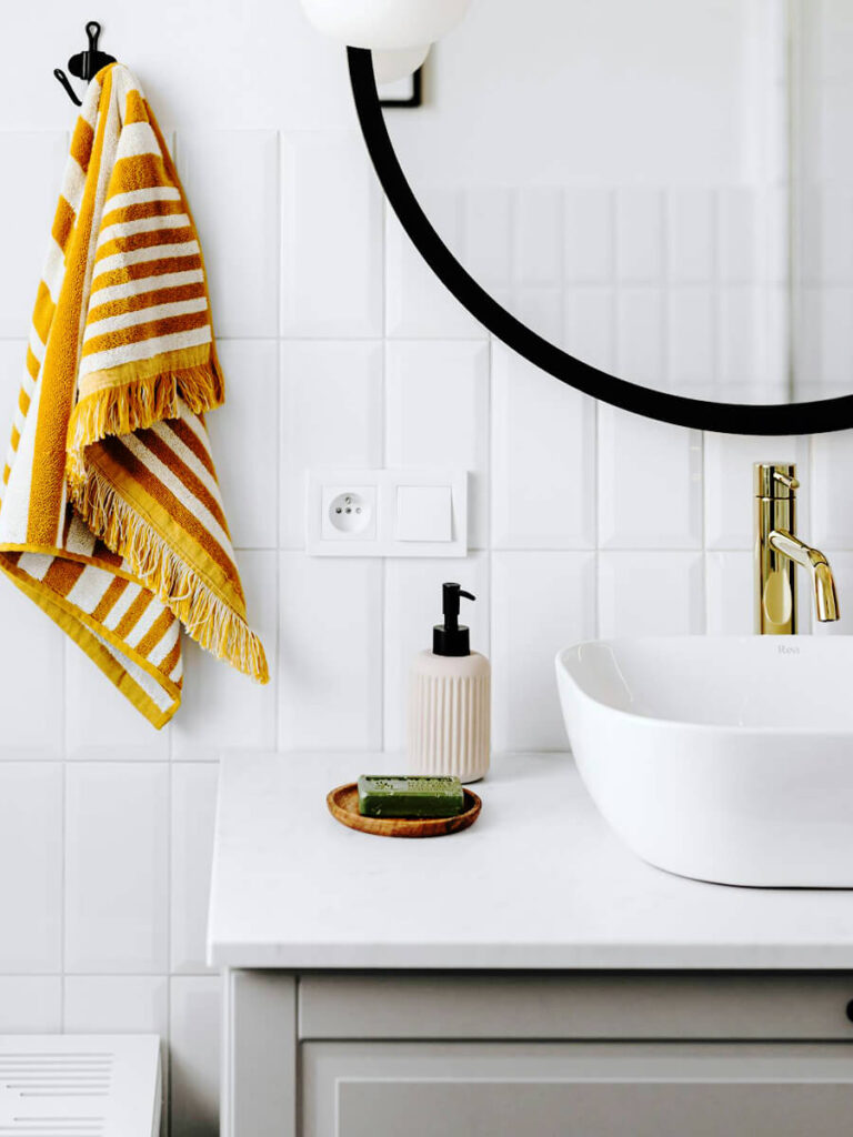 Bathroom with a white porcelain sink, wall-mounted round mirror, and matte white beveled subway tiles laid in a vertical grid, accented by a towel and soap dispenser for a crisp, modern fixture area.