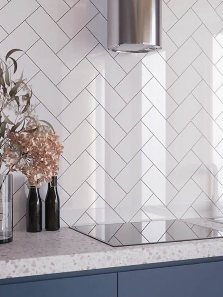 Modern kitchen backsplash of glossy white subway tiles set in a precise herringbone pattern with crisp dark grout, accompanying terrazzo countertop, blue cabinets, and chrome vent hood for an elegant, bright style.