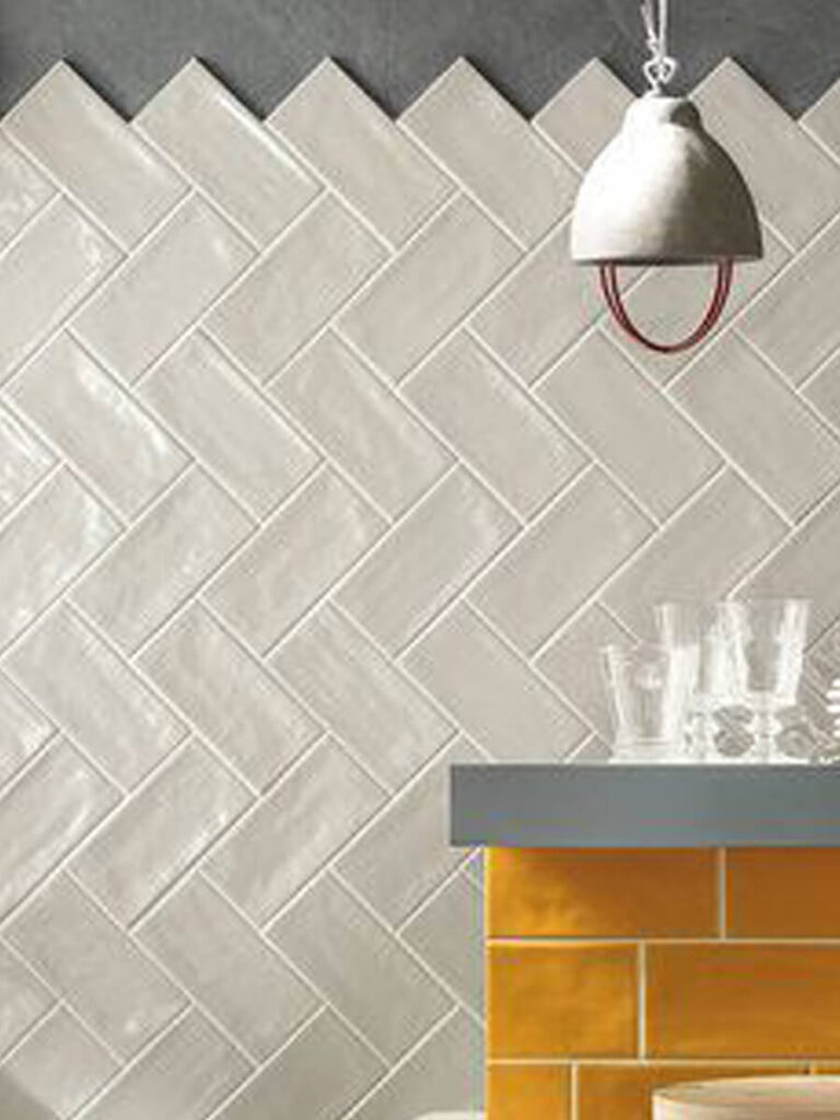 Kitchen backsplash with matte white subway tiles arranged in a herringbone pattern, finished in a zigzag edge at the top, contrasted by a lower bar finished in glossy mustard yellow stacked tiles, and a concrete pendant lamp above.