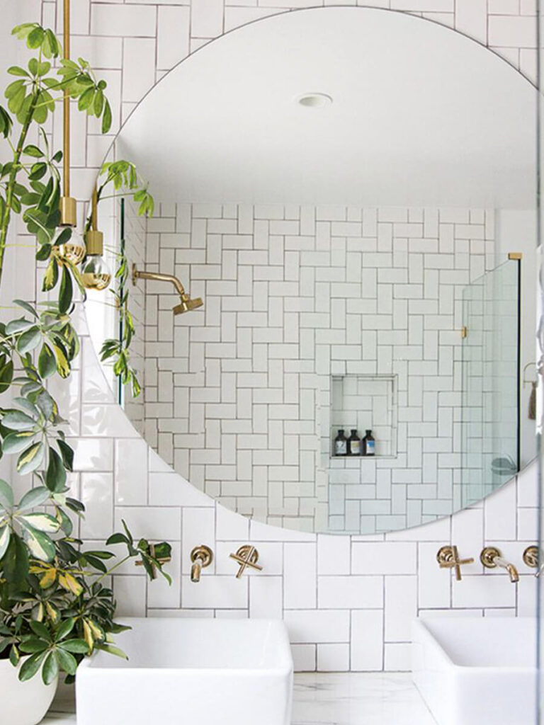Chic bathroom featuring matte white subway tiles in a vertical herringbone layout, spotted in a large round mirror above double sinks, with brass hardware and tall green plants for a spa-like vibe.