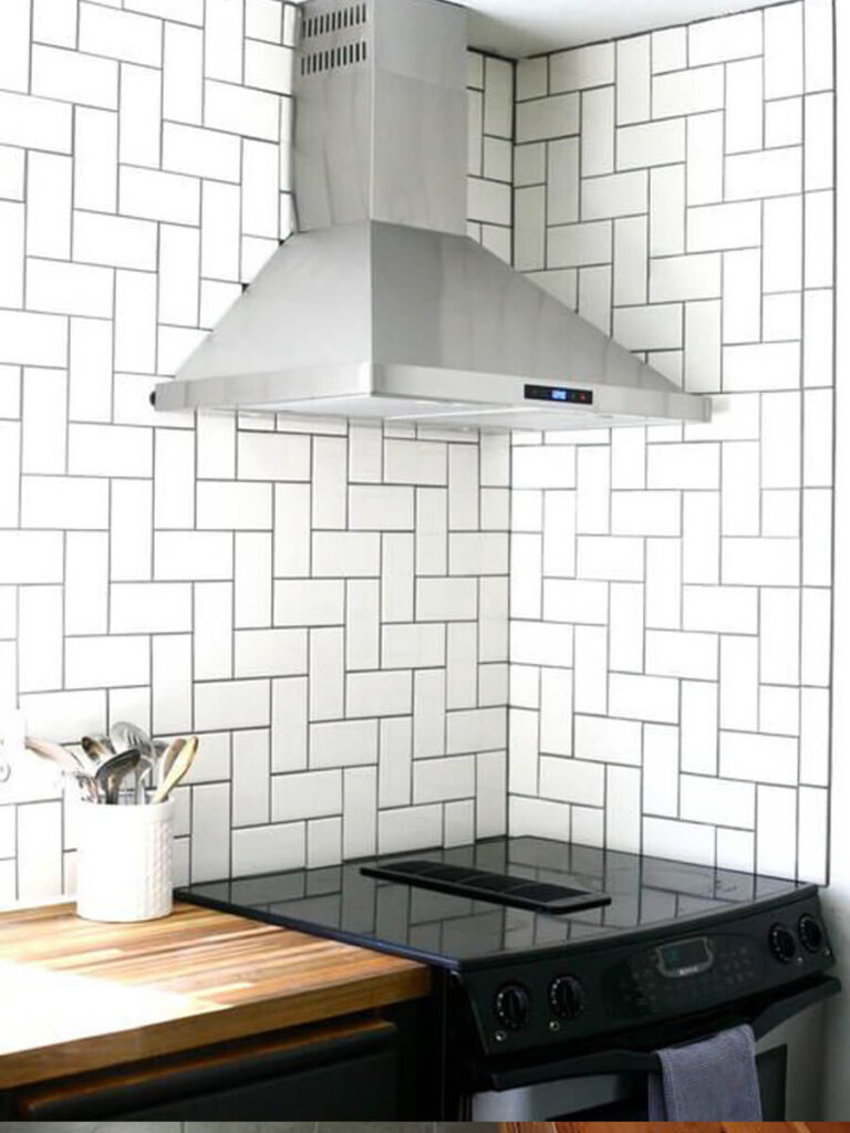 Kitchen backsplash with glossy white tiles laid in a vertical herringbone pattern, set behind a modern electric stove, with kitchen utensils and accessories for a sleek, cook-friendly environment.