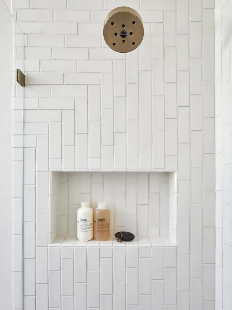 Minimalist bathroom shower wall with matte white subway tiles laid in a vertical herringbone pattern and a built-in shelf for toiletries, centered below a round brass showerhead.