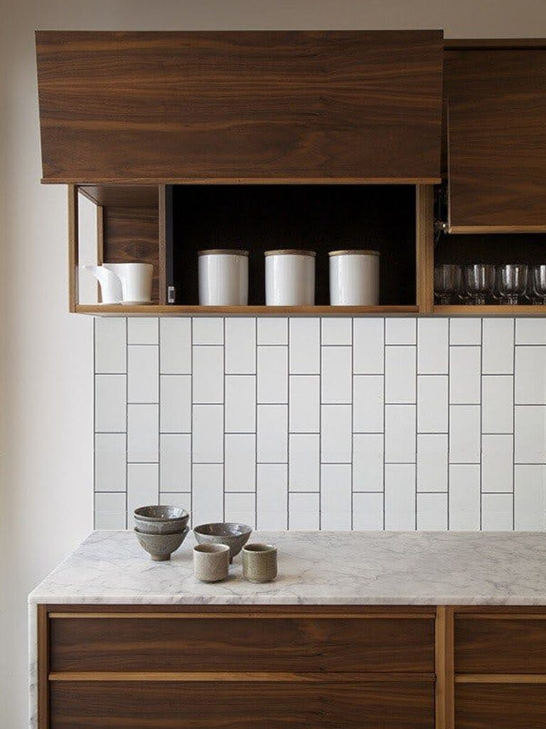 Kitchen backsplash with glossy white subway tiles laid in a classic vertical running bond, paired with dark wood cabinets, open shelf, ceramic canisters, and a marble countertop.