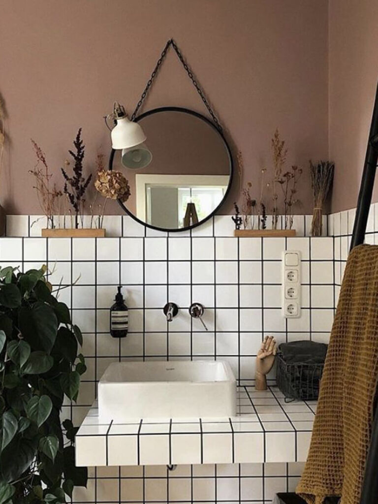 Contemporary bathroom design with white square tiles, black grout, soft taupe painted walls, a hanging round mirror, and dried botanicals for decoration.