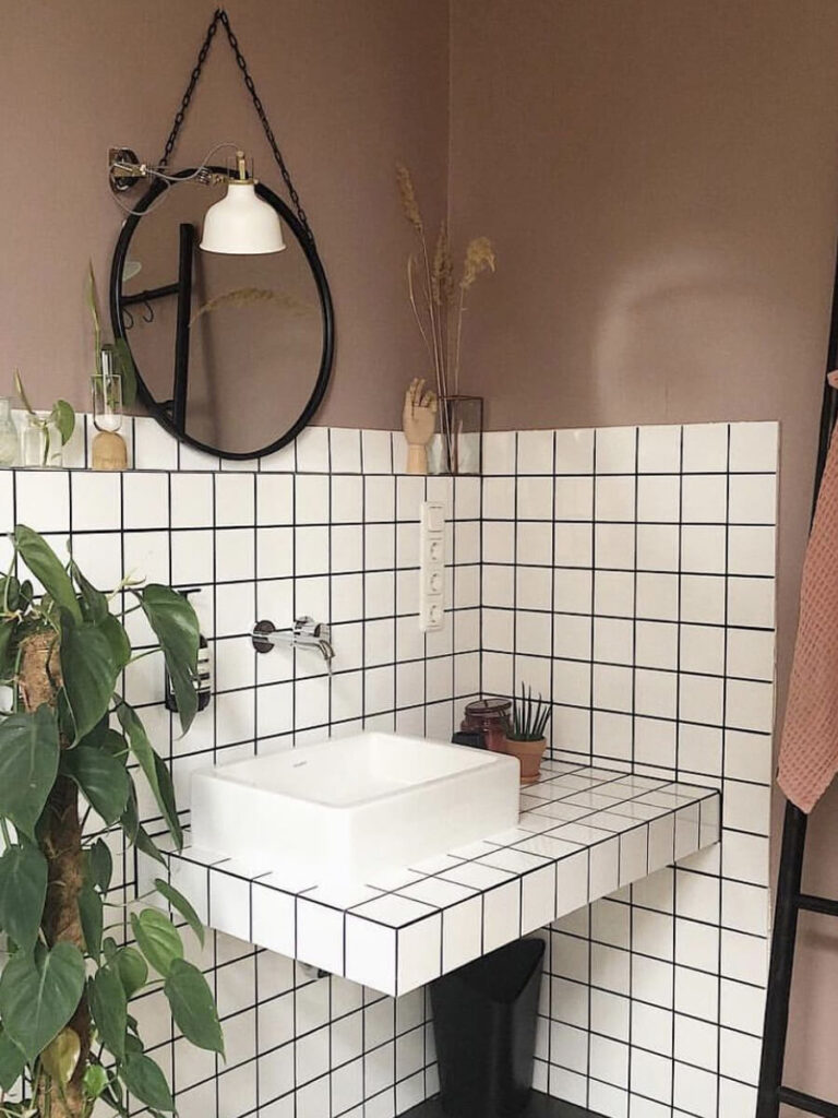 Bathroom with square white tiles and black grout, a round mirror hanging by a chain, matte taupe-pink wall paint, and botanical accents.