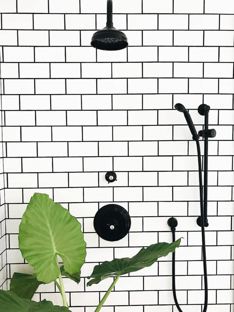 Black shower fixtures on a wall of horizontally laid white subway tiles with dark grout, accented by large green tropical leaves in the foreground.