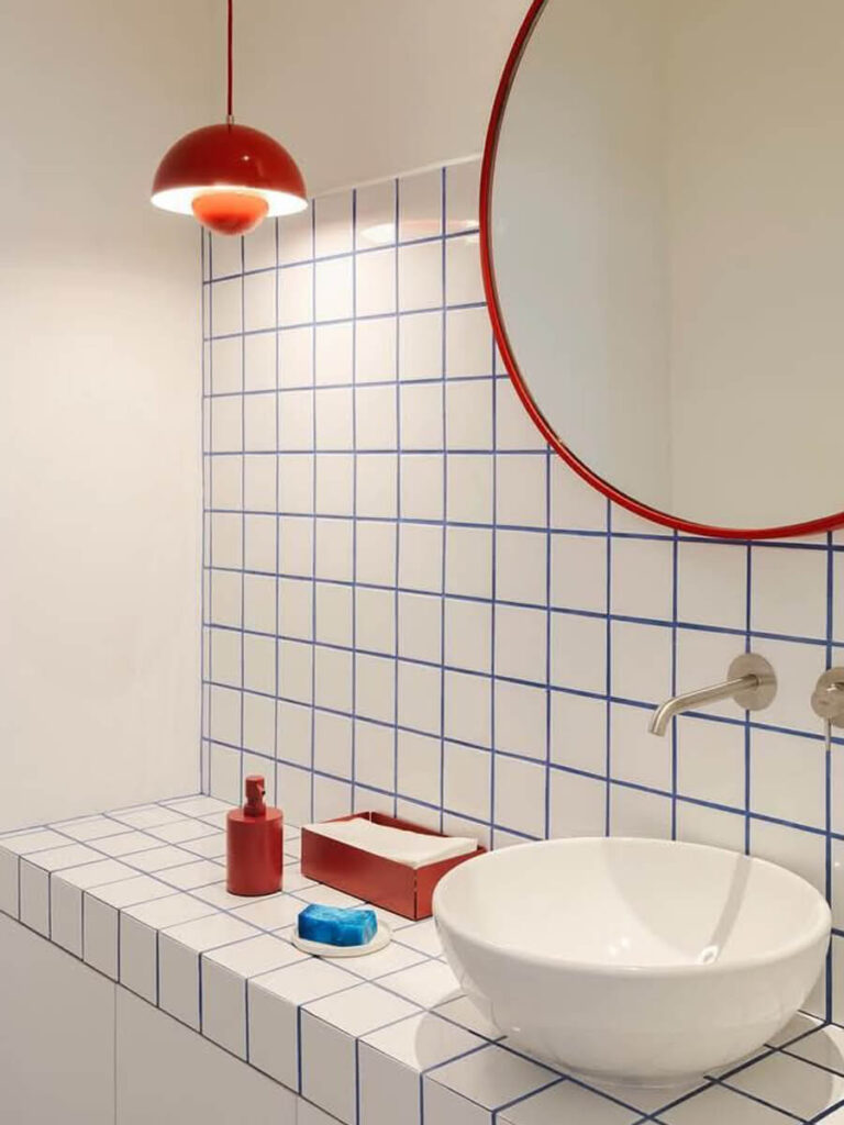 Minimalist bathroom with white square tiles outlined in blue grout, a round mirror with a red frame, and a red pendant lamp for a pop of color.